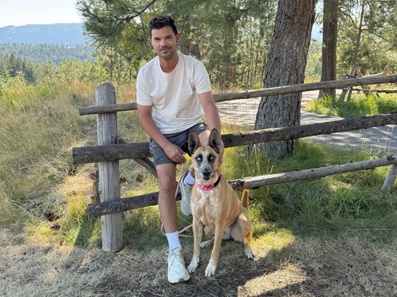 Person in white t-shirt, gray shorts, and white sneakers sitting on a wooden fence outdoors; face obscured. A dog with black facial markings and a red collar sits beside them, looking at the camera. Trees and mountains in the background.