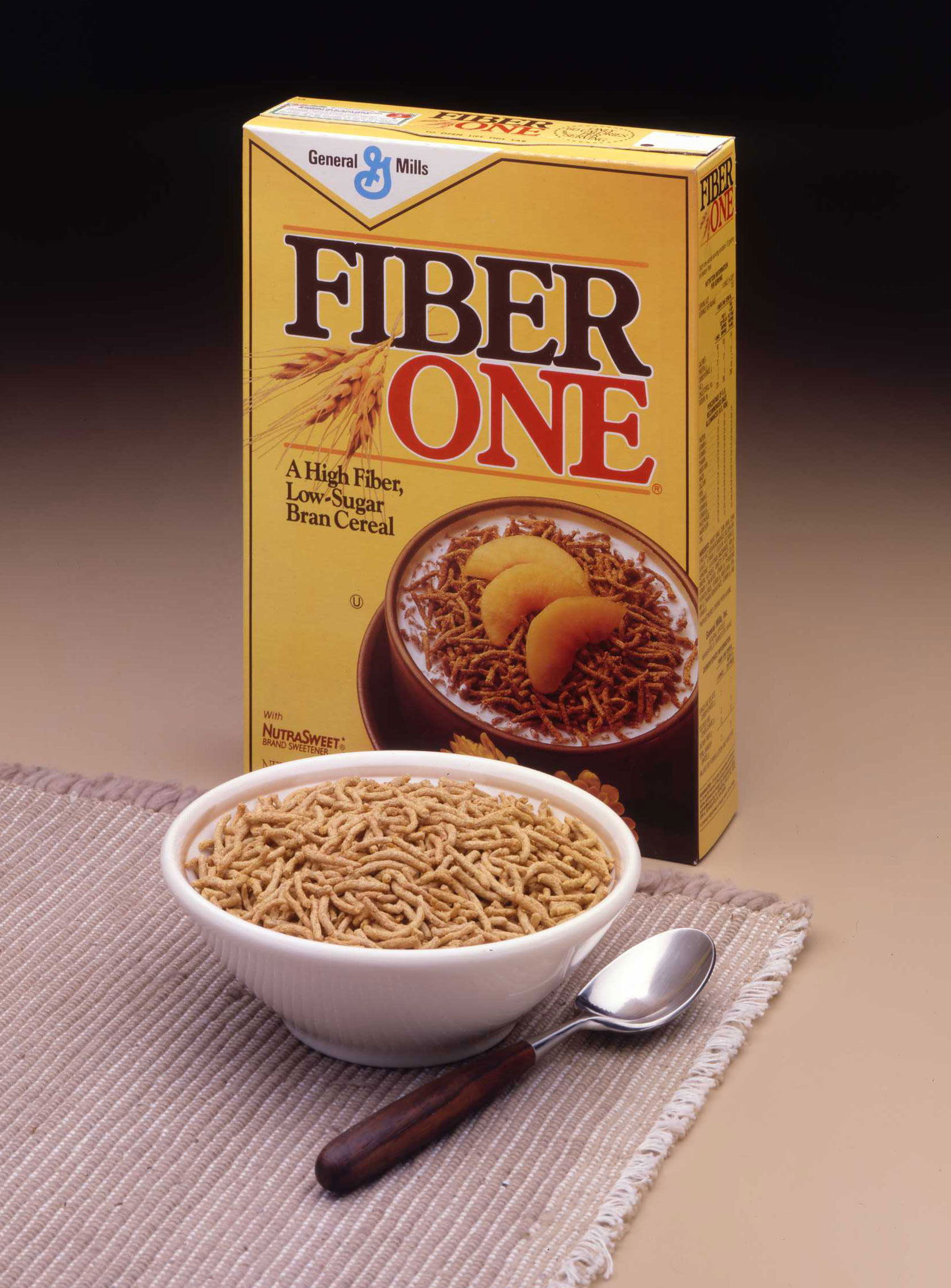 A classic product shot showing a yellow box of original Fiber One bran cereal. In front of the box, there is a full white bowl of the cereal placed on a textured placemat, with a spoon resting next to it.